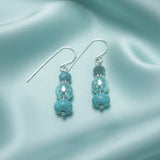 A pair of dangling earrings with silver-tone fishhook wires and three turquoise-blue beads on each earring, placed on a beige textured surface.